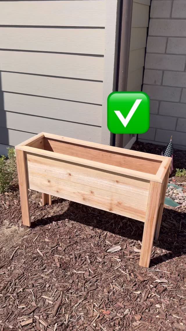 Rectangular light wood planter box with legs sitting on brown mulch outdoors.