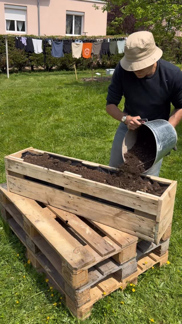Man filling a long wooden pallet crate planter with soil in a garden.