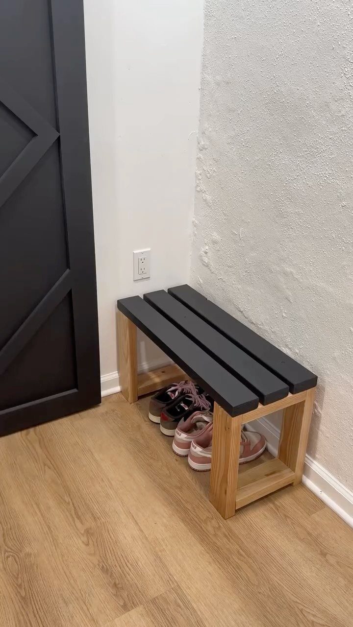 Wooden entry bench with black slatted top and shoes stored underneath against white wall.