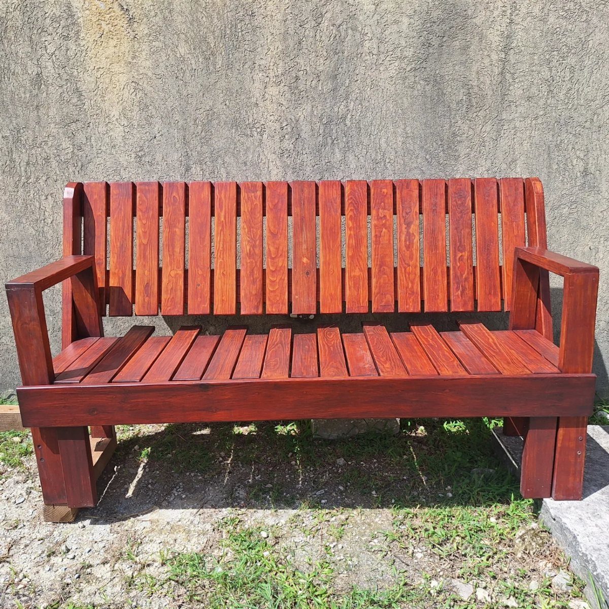14 Woodworking Ideas to Make Money From Your Shop 14 Reddish-brown wooden garden bench with slatted back against a grey concrete wall.