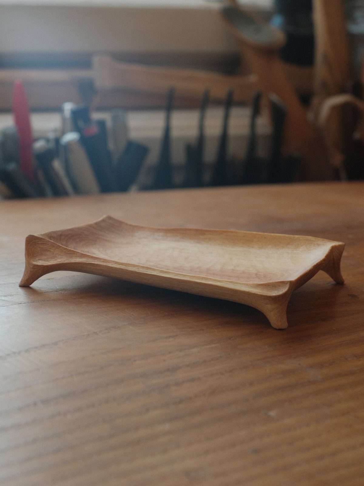 14 Woodworking Ideas to Make Money From Your Shop 10 Hand-carved light brown wooden tray resting on a wooden workbench with tools behind.